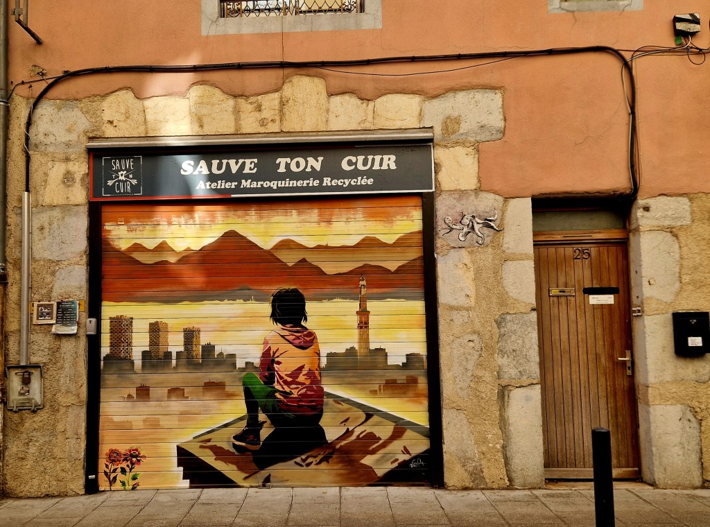 Ein kunstvoll bemalter Rolladen in Grenoble zeigt eine nachdenkliche Figur zwischen Stadt und Bergen – ein kleines Kunstwerk mitten in der Altstadt.
