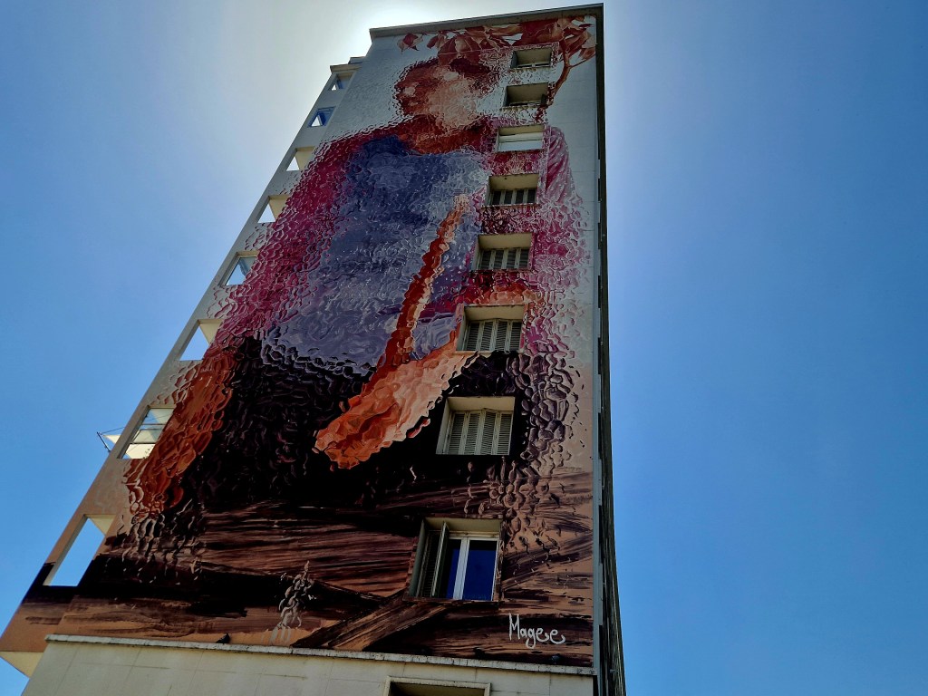 Riesiges Street-Art-Kunstwerk an einer Hausfassade in Grenoble, das eine Person vor blauem Himmel zeigt.